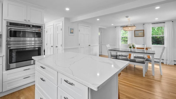 A modern kitchen with white cabinetry, a large island, and a dining area with a glass table and chairs. - Stunning Cape Home Near Chatham (Brewster)