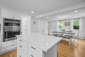 A modern kitchen with white cabinetry, a large island, and a dining area with a glass table and chairs. - Stunning Cape Home Near Chatham (Brewster)