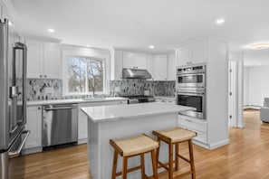 Una cocina moderna con muebles blancos, una isla grande con taburetes, lavavajillas, estufa y horno.