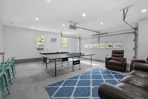 Una habitación con mesa de ping pong, sillas verdes, sofá de cuero marrón y un proyector montado en el techo.