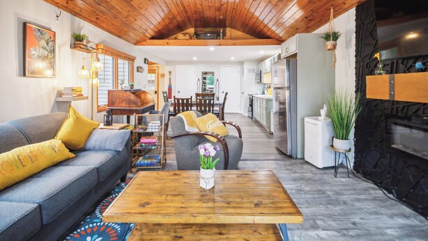 Ein gemütliches Wohnzimmer mit Holzboden, einer Couch mit gelben Kissen, einem Couchtisch mit Blumen und einer Küche im Hintergrund.