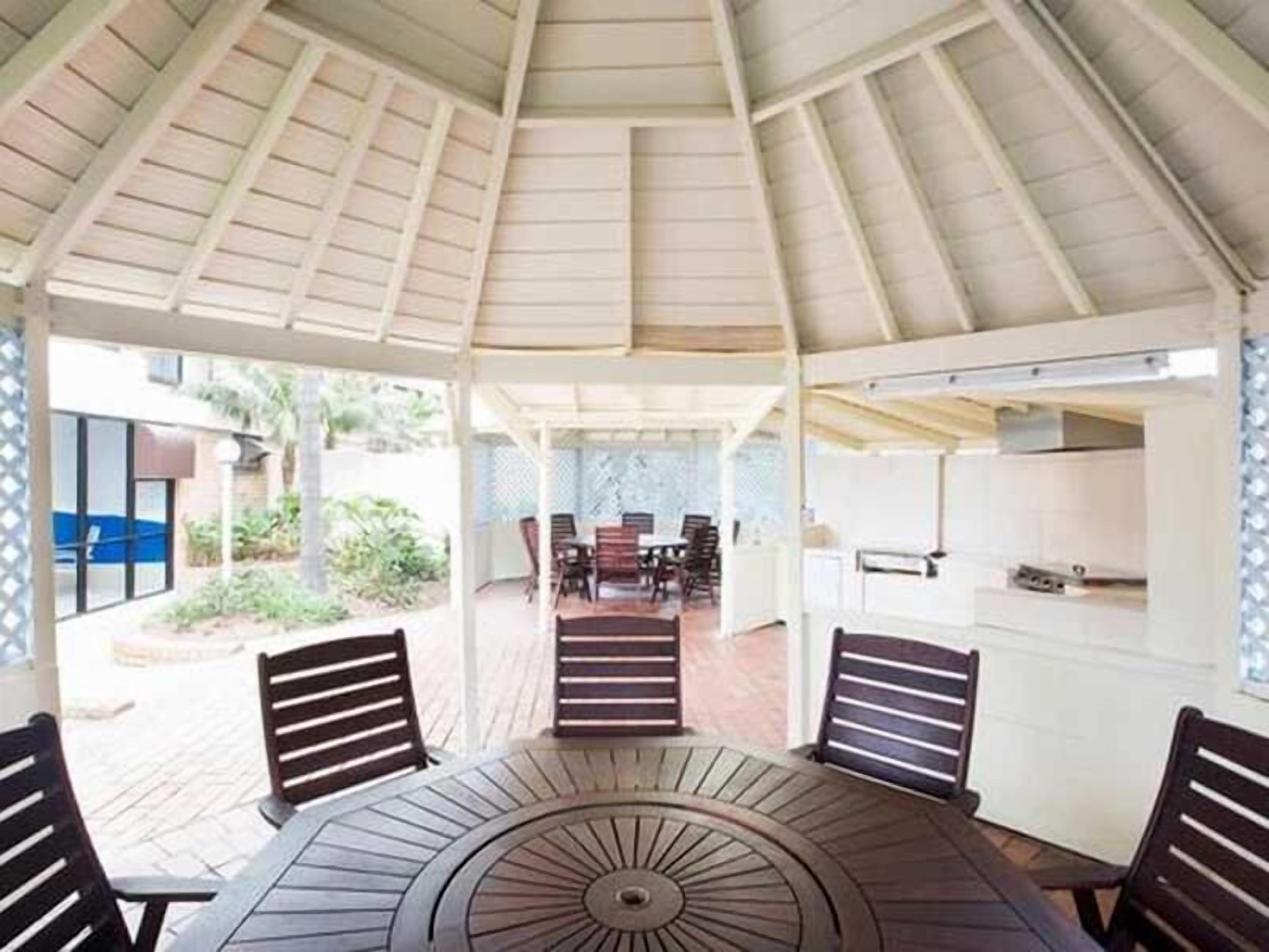 A covered outdoor dining area with a round table and chairs, a kitchen area in the background, and a view of the surrounding garden.