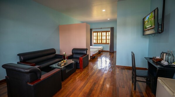 A hotel room with a sofa, a small table with books, a bed, a TV, and a window with curtains.