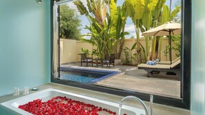 Une salle de bain avec une baignoire remplie de pétales de rose, une fenêtre donnant sur un espace piscine avec des chaises et un parasol.