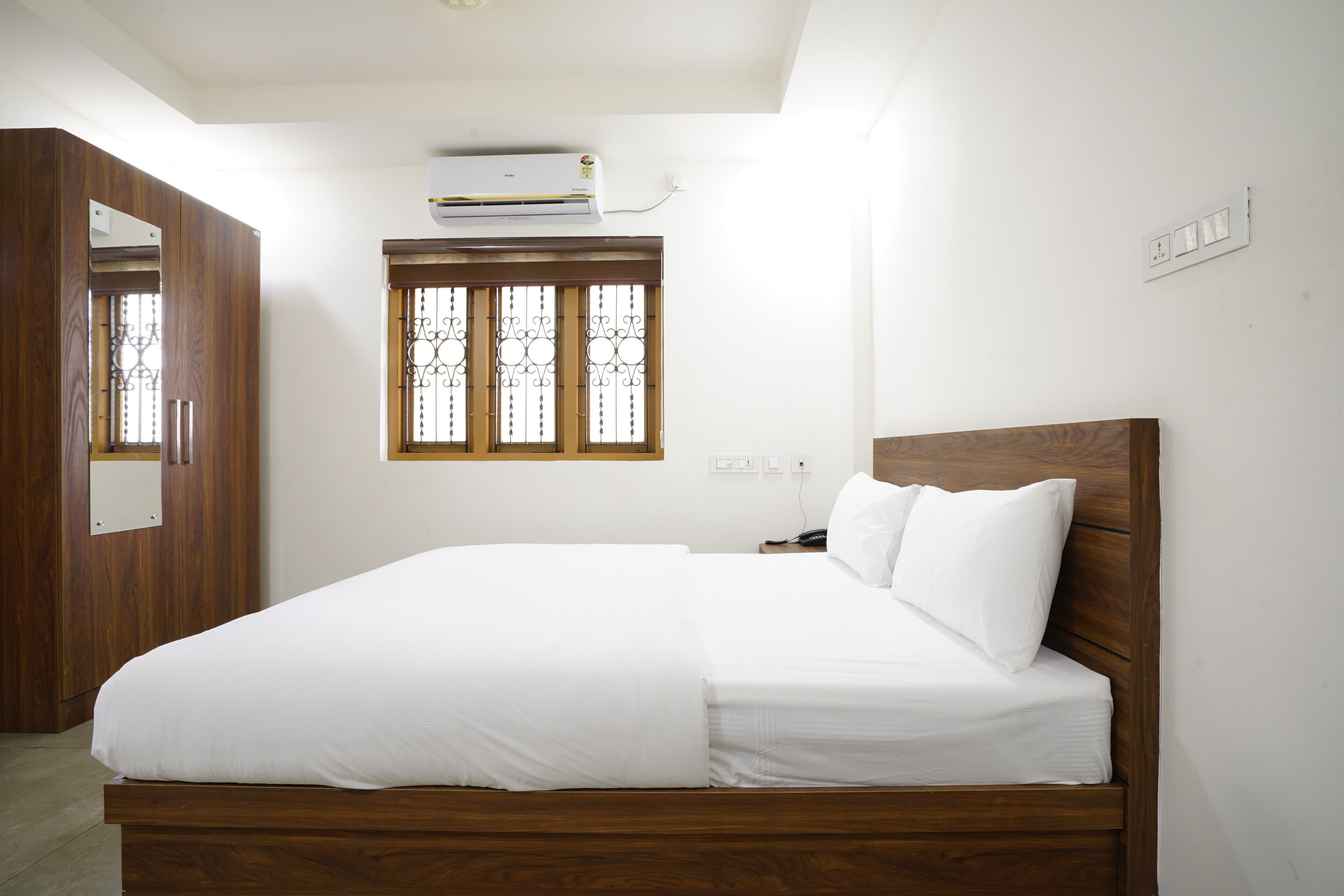 A bedroom with a wooden bed, white bedding, a window with decorative bars, an air conditioning unit, and a wardrobe.