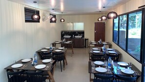 A dining area with tables set for meals, a kitchen in the background, and large windows providing natural light. - Halo Residency (Kanayannur)