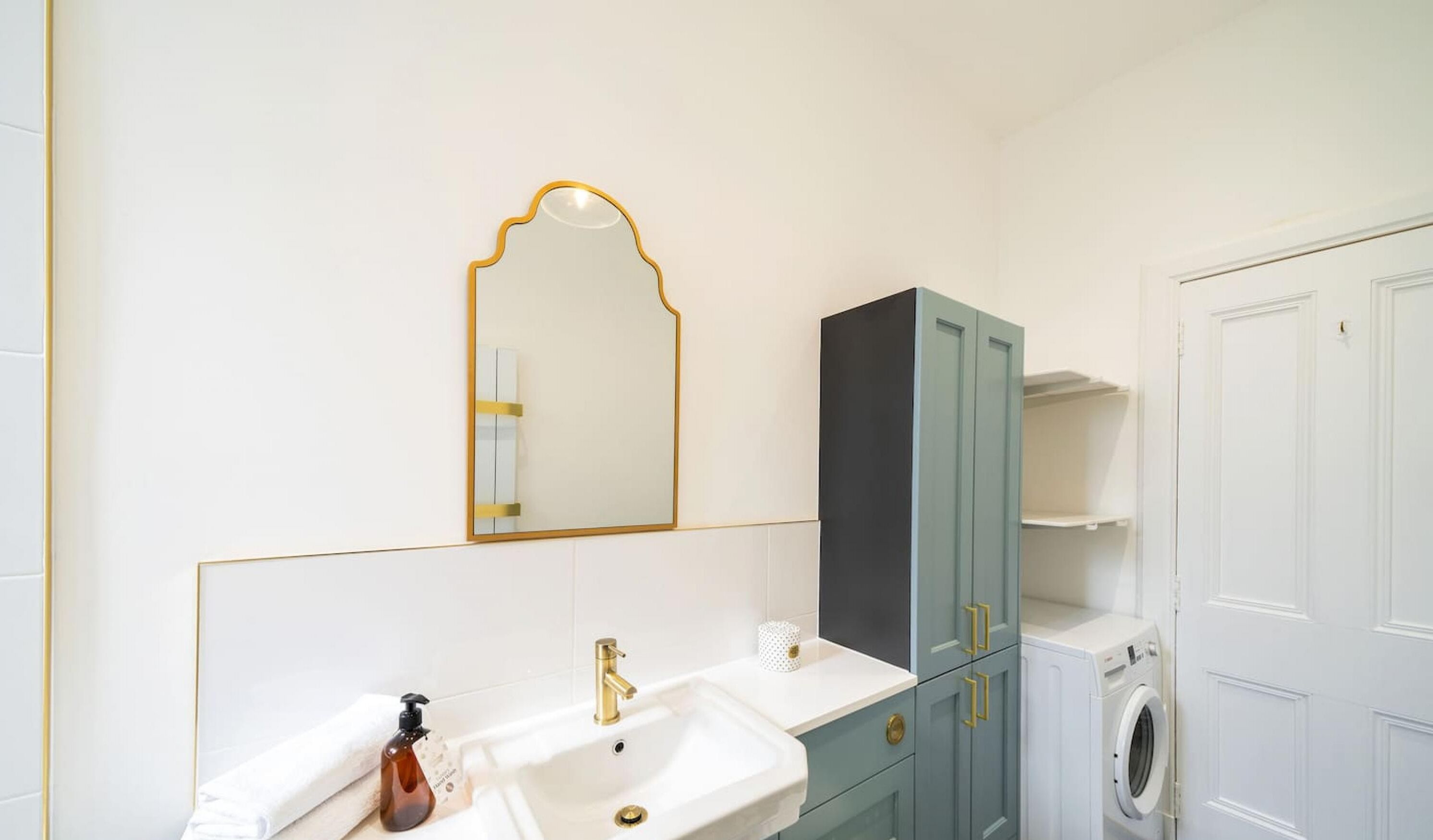A bathroom with a white sink, gold faucet, a mirror with a gold frame, a teal cabinet, and a washing machine.