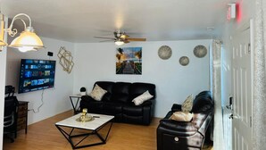 Ein Wohnzimmer mit schwarzem Ledersofa, einem Couchtisch, einem an der Wand befestigten Fernseher und einem Deckenventilator.