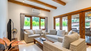 A spacious living area with wicker furniture, large windows, and a view of greenery outside. - Peaceful 3BR Villa Located in Pecatu - 10 Min to the Beach (Pecatu)