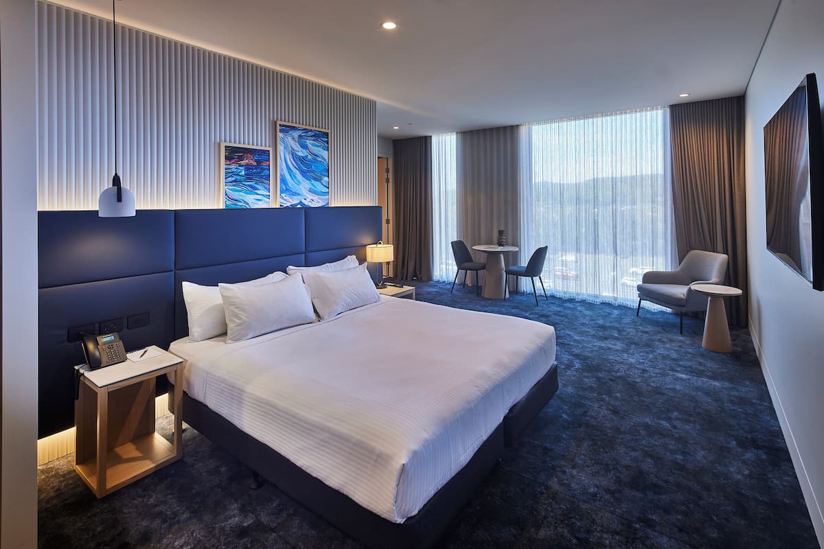 A modern hotel room with a large bed, a bedside phone, a small table, a chair, and a view of the city through large windows.