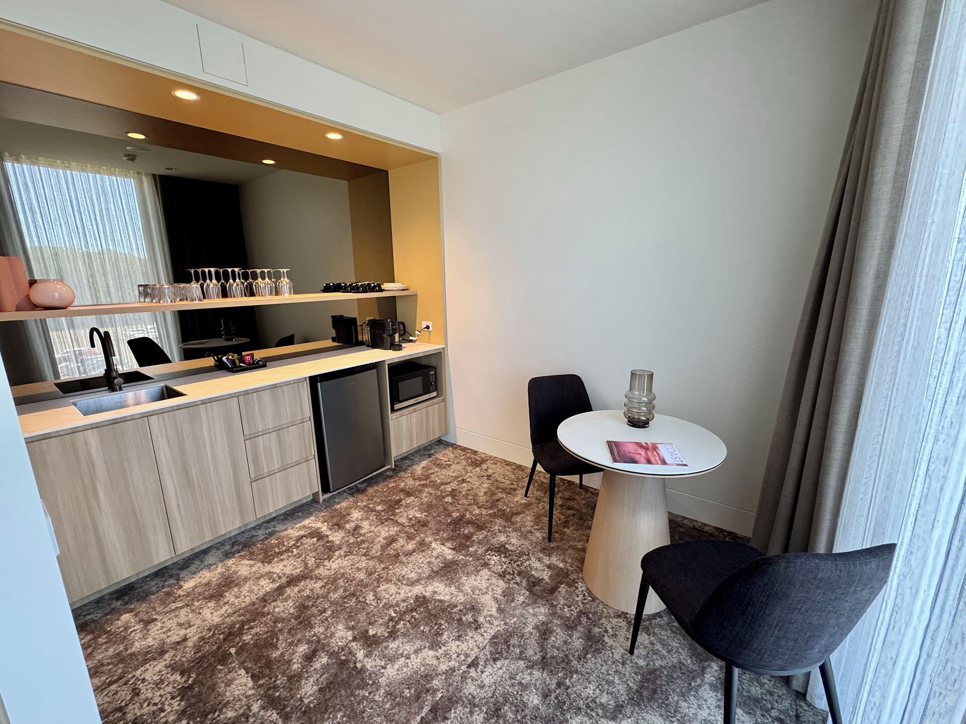 A compact kitchenette with a sink, coffee maker, and microwave. There is a small round table with two chairs and a glass vase.