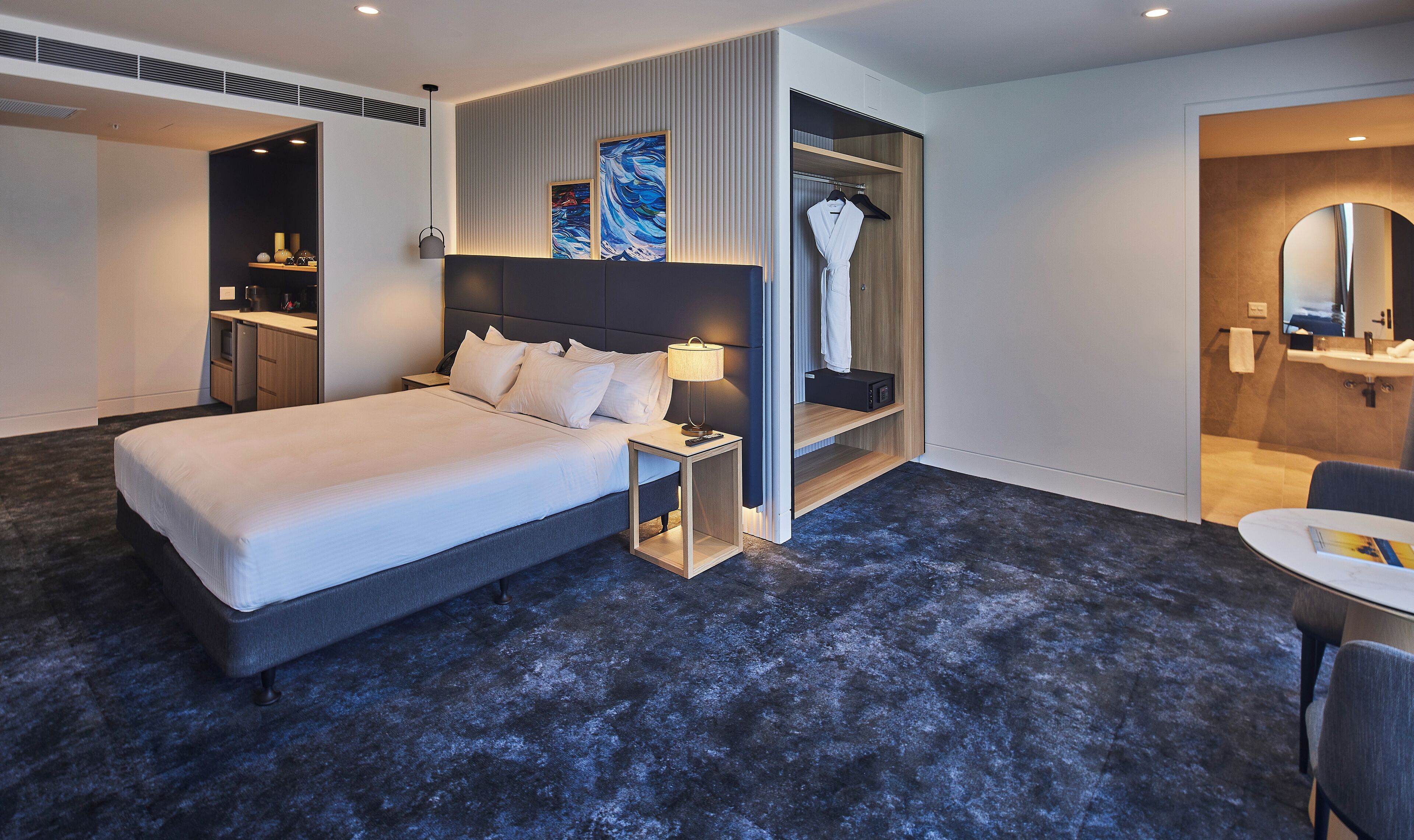 A modern hotel room with a large bed, a bedside table with a lamp, a wardrobe, and a bathroom with a mirror and sink.
