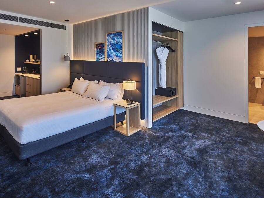 Una habitación de hotel moderna con una cama grande, una mesita de noche con lámpara, un armario y un baño con espejo y lavamanos.