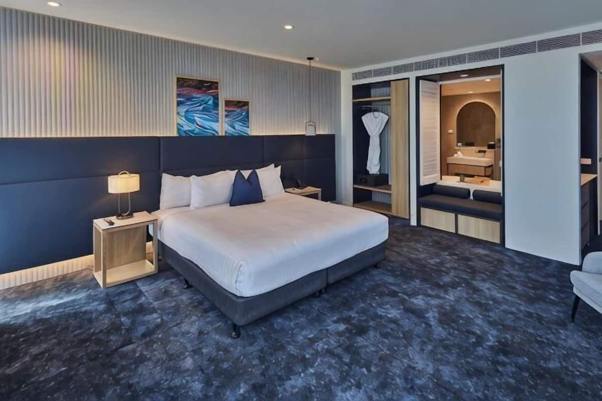 A modern hotel room with a large bed, bedside tables, a wardrobe, and a bathroom visible through an open door.