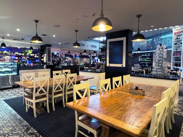 Un restaurante con mesas y sillas de madera, lámparas colgantes y una zona de mostrador con una máquina de café.