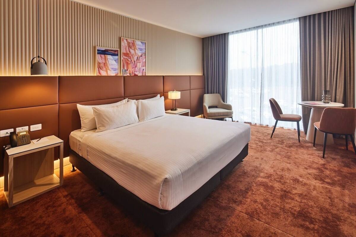 A hotel room with a large bed, a bedside table with a lamp, a chair, a small table, and a view of the city through large windows.