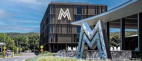A modern building with a prominent 'M' logo, a landscaped area with a water feature, and a clear sky.