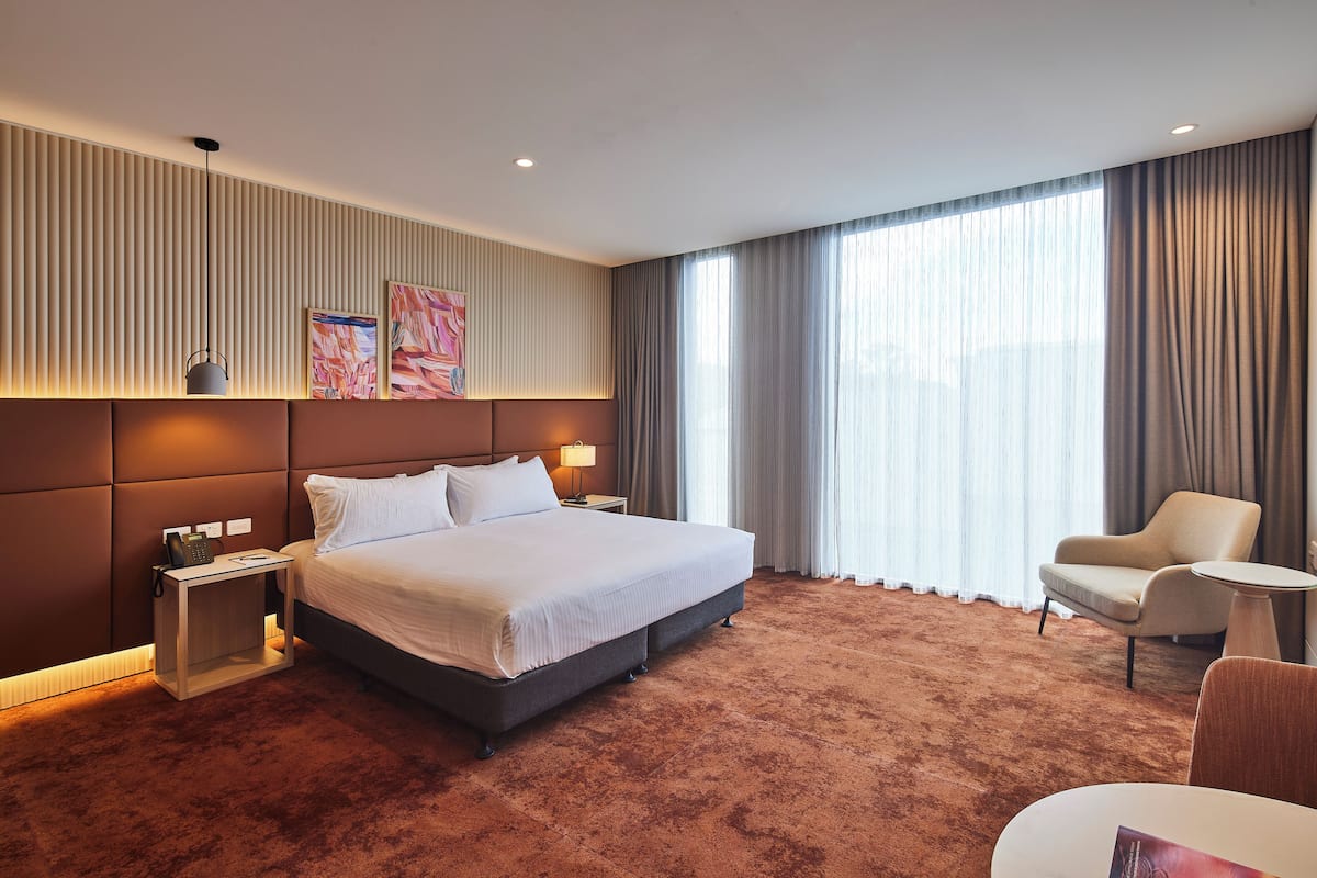 A modern hotel room with a large bed, a bedside table with a lamp, a chair, and a large window with curtains.