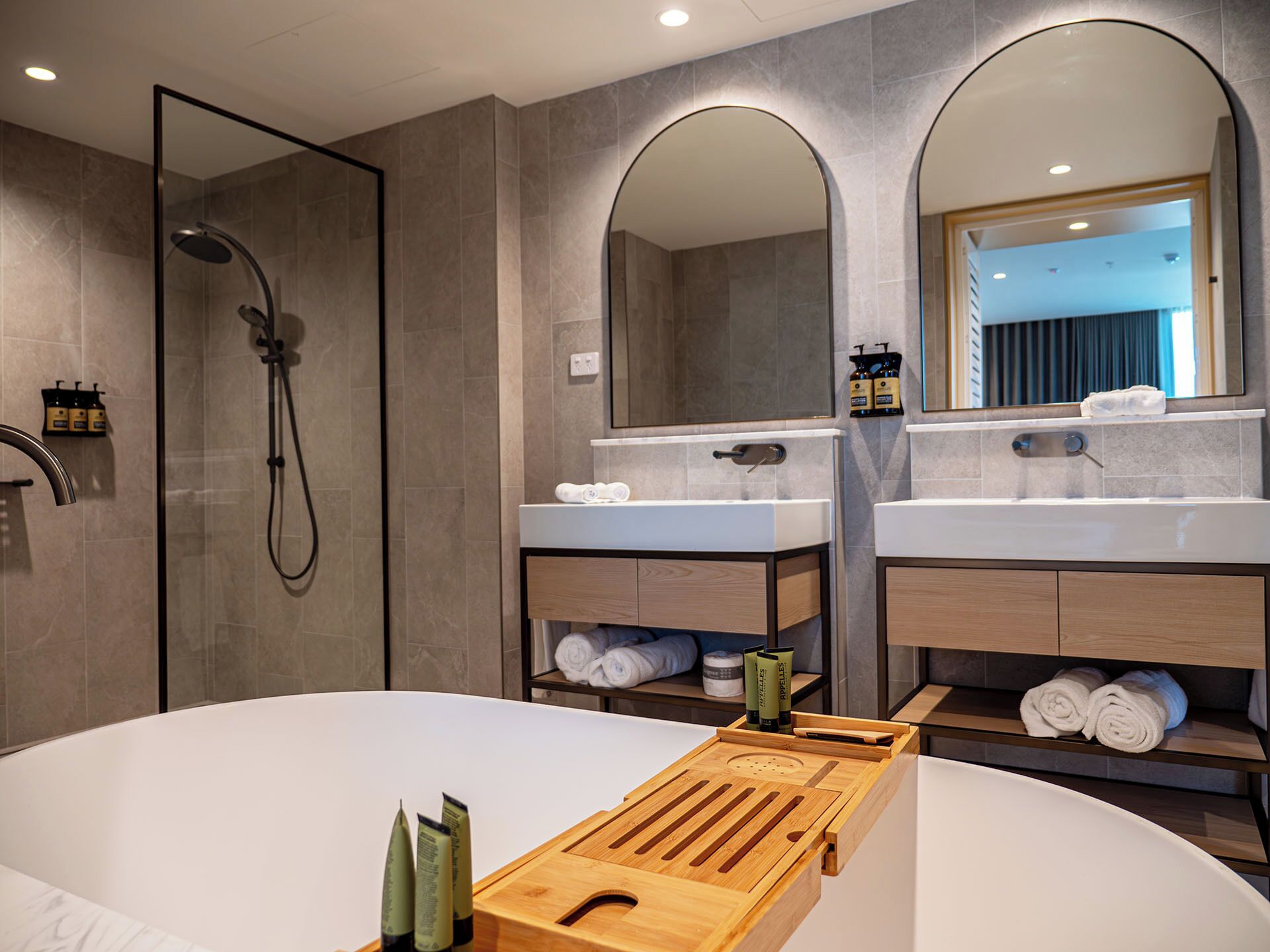 A modern bathroom with a shower, two sinks, a large mirror, and a wooden tray with toiletries.