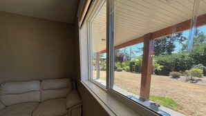A room with a beige sofa against a wall, a large window with a view of a garden and outdoor seating area.