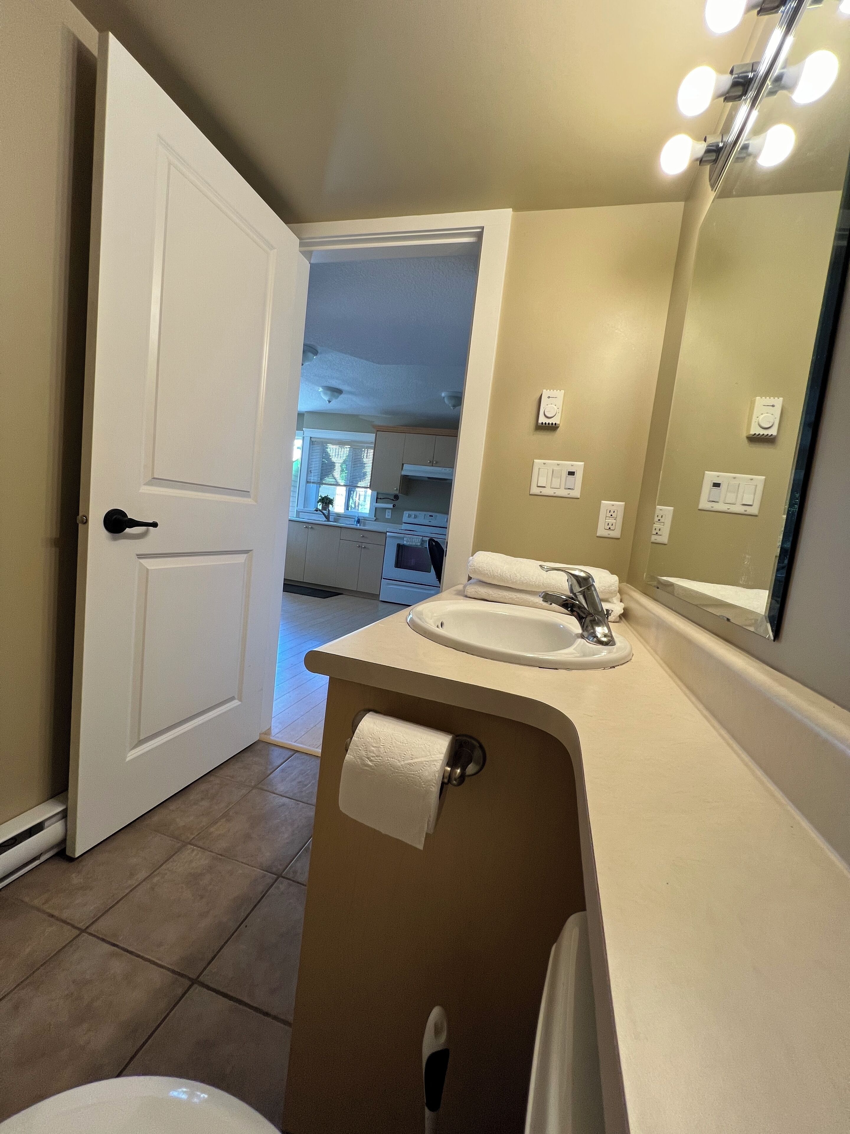 A bathroom with a sink, mirror, and toilet. There is a door leading to a kitchen area.