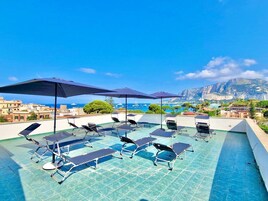 Piscina no terraço com espreguiçadeiras e guarda-sóis, com vista para uma cidade costeira e uma montanha.