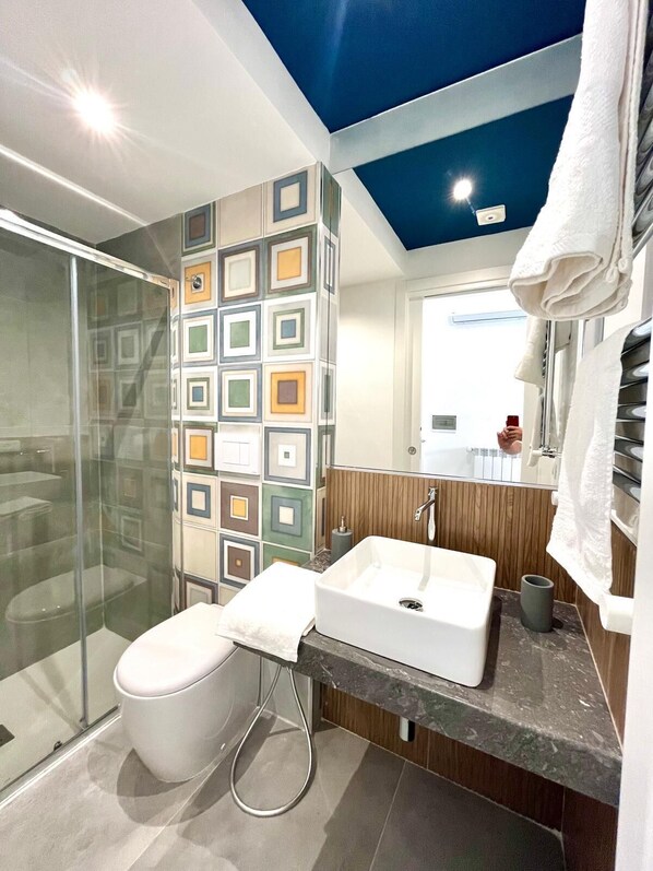 A modern bathroom with a glass shower enclosure, a white sink, and a tiled wall with a geometric pattern. - Mongibello rooftop (Palermo)