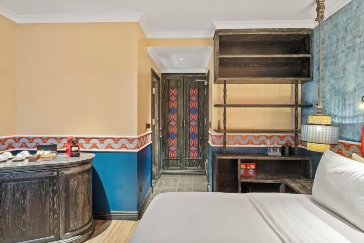 A hotel room with a bed, a round wooden side table, a shelf with decorative items, and a patterned door.