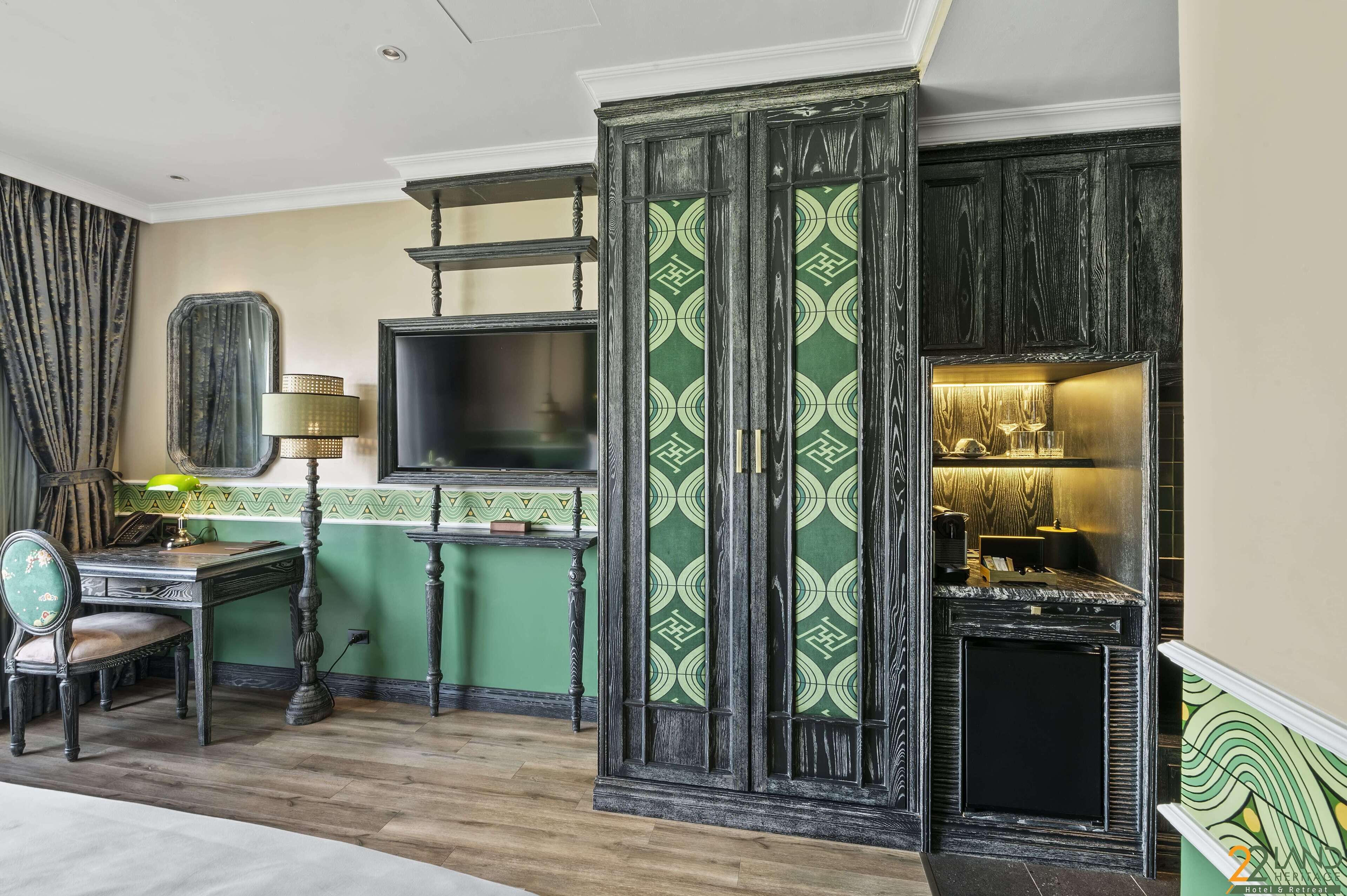 A room with a green patterned wall, a wooden cabinet with a television, a desk with a lamp, and a fireplace.