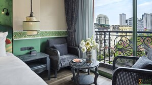 Une chambre d’hôtel avec un lit, une petite table ornée d’un vase de fleurs, une chaise et un balcon offrant une vue sur la ville.