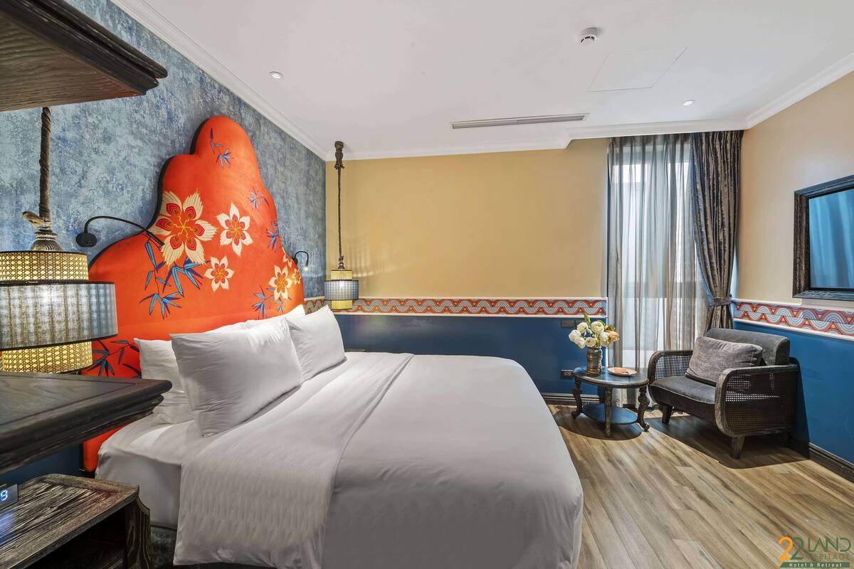 A hotel room with a large bed, a small table with a lamp, a chair, and a window with curtains.