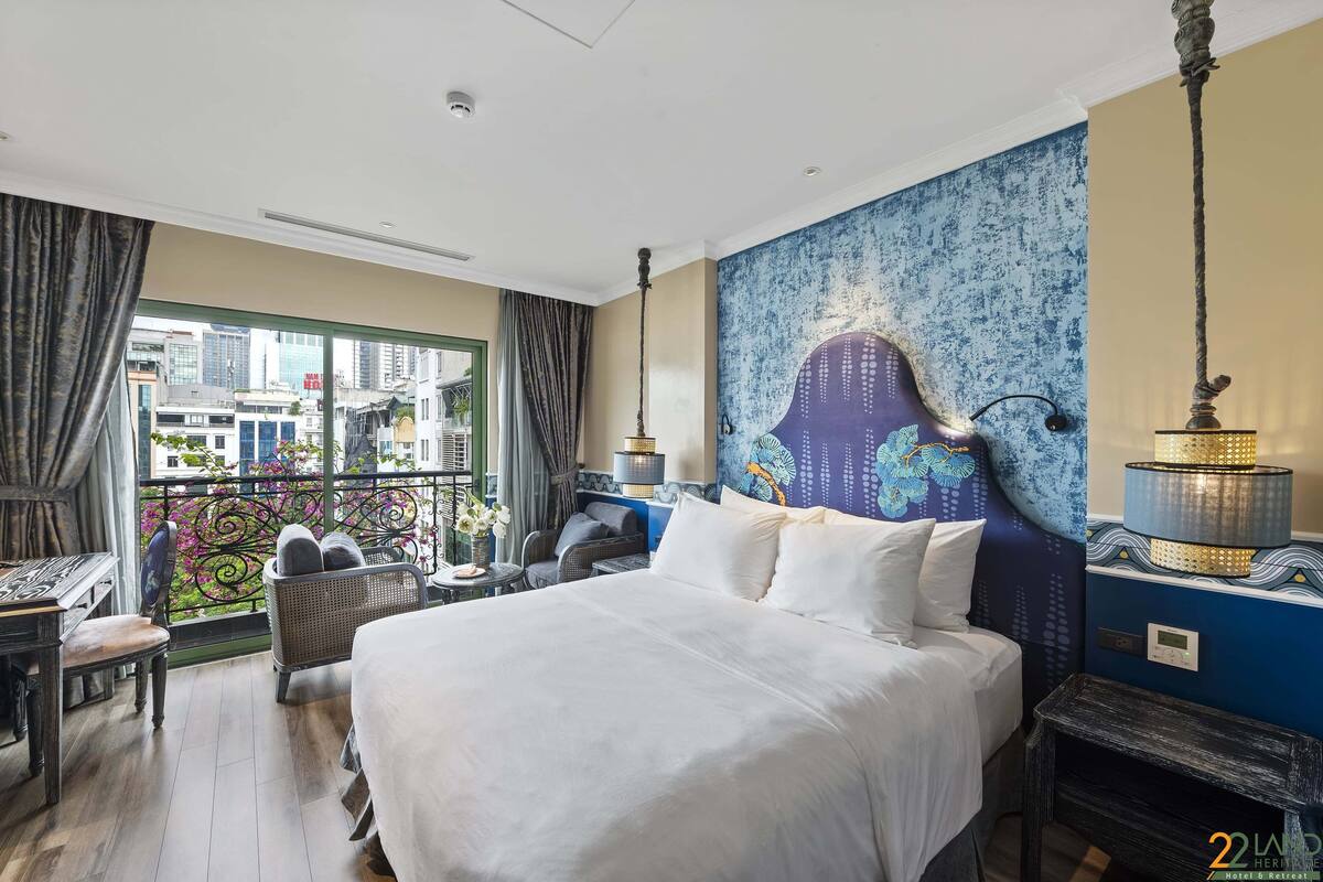 A hotel room with a large bed, a desk, a chair, a small table, and a balcony with a view of the city.