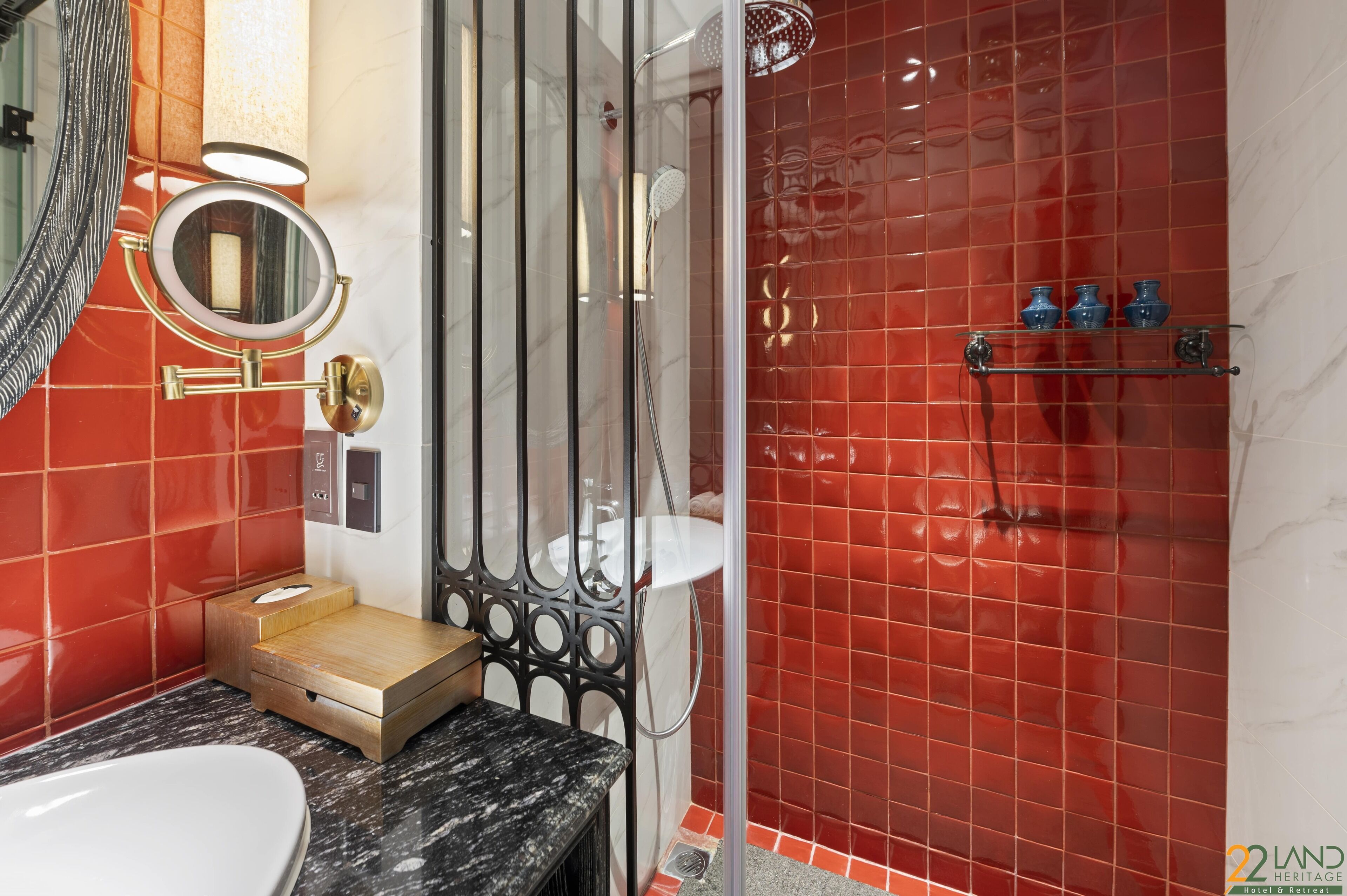 A bathroom with a red tiled shower, a wooden vanity, a white sink, and a mirror with a round frame.