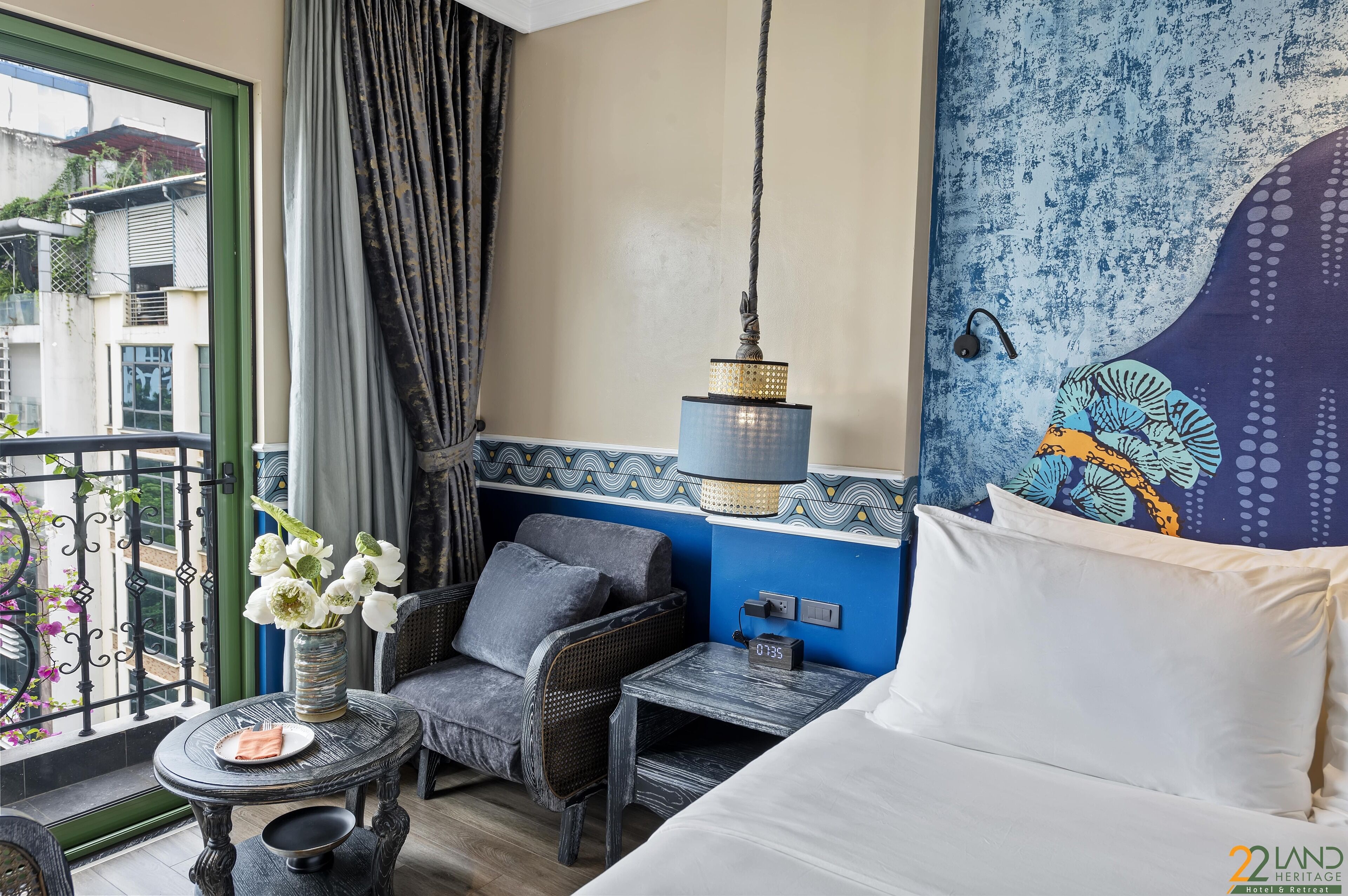 A hotel room with a bed, a small table with a vase of flowers, a chair, a hanging light fixture, and a view of the outside balcony.