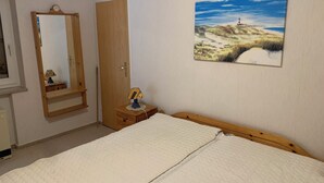 Un dormitorio con cama, cabecera de madera, mesita de noche con lámpara, puerta y un cuadro de un paisaje.