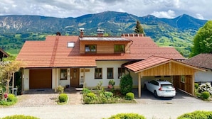 Ein zweistöckiges Haus mit rotem Ziegeldach, einer Garage und einem darin geparkten Auto. Das Haus wird von üppiger Vegetation und Bergen im Hintergrund umgeben.
