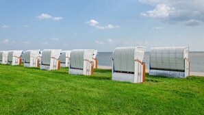 Eine Reihe weißer Korbstühle mit braunen Sitzflächen und Rückenlehnen, auf einer Rasenfläche am Meer aufgereiht.