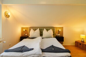 A double bed with white linens, two bedside tables with lamps, a headboard with vertical slats, and a small wooden nightstand with a lamp.