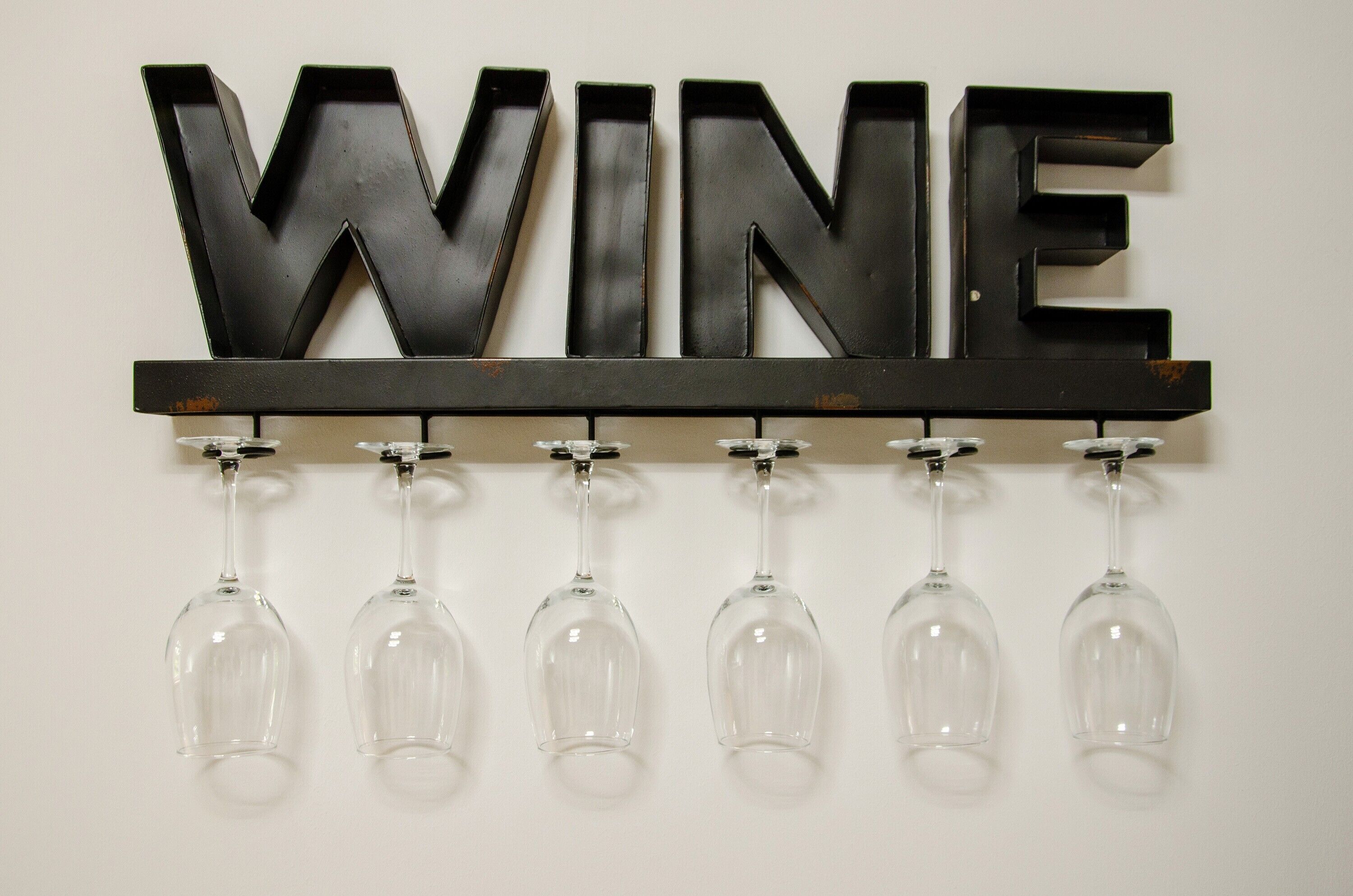 Ein an der Wand befestigtes Schild mit dem Wort 'WINE' und darunter sechs hängende Weingläser.