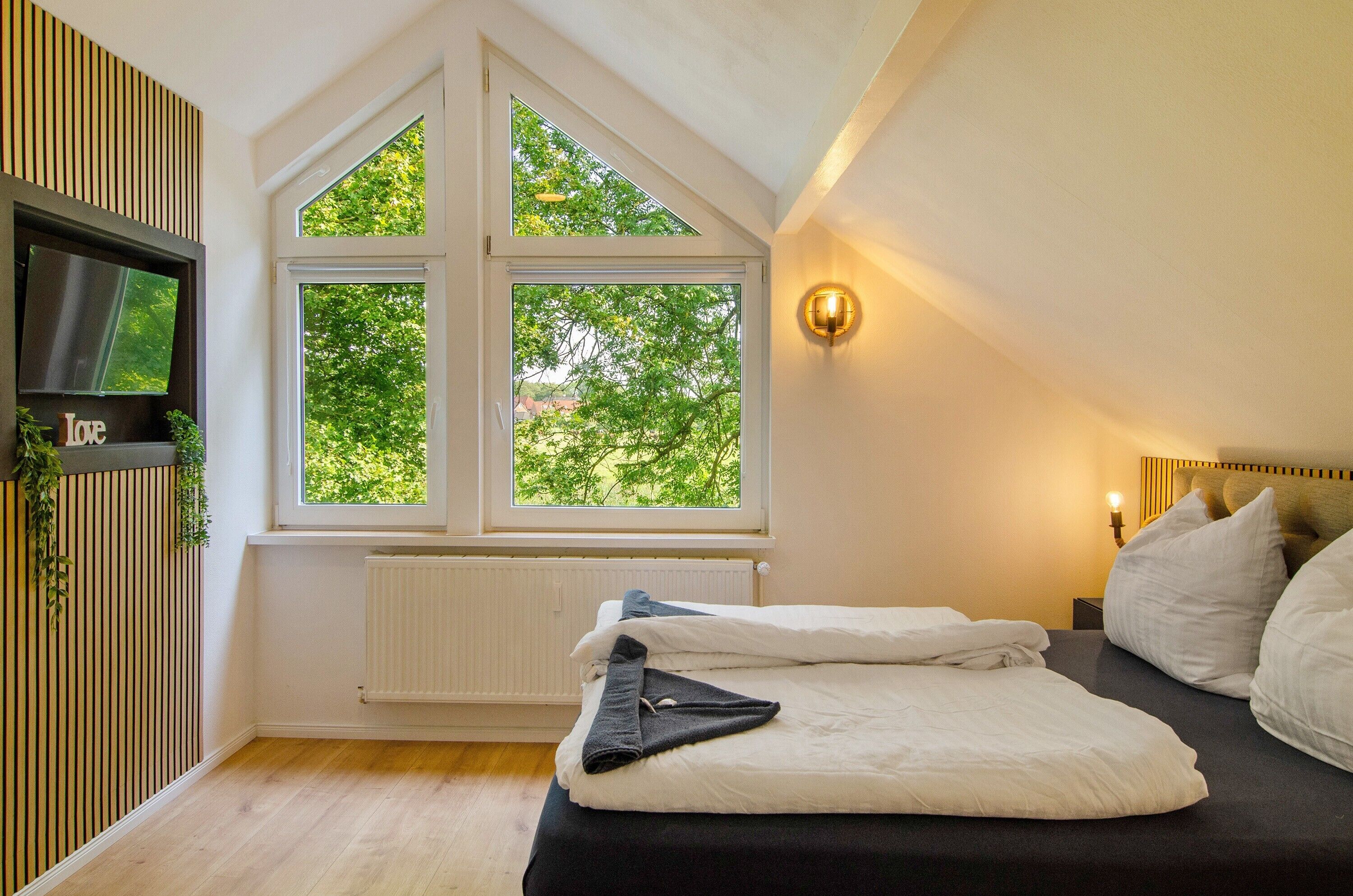 Ein Schlafzimmer mit einem Bett, einem an der Wand befestigten Fernseher, einem Fenster mit Blick auf Bäume und einer Wandleuchte.