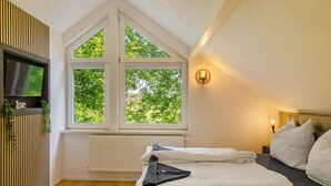 Ein Schlafzimmer mit einem Bett, einem an der Wand befestigten Fernseher, einem Fenster mit Blick auf Bäume und einer Wandleuchte.