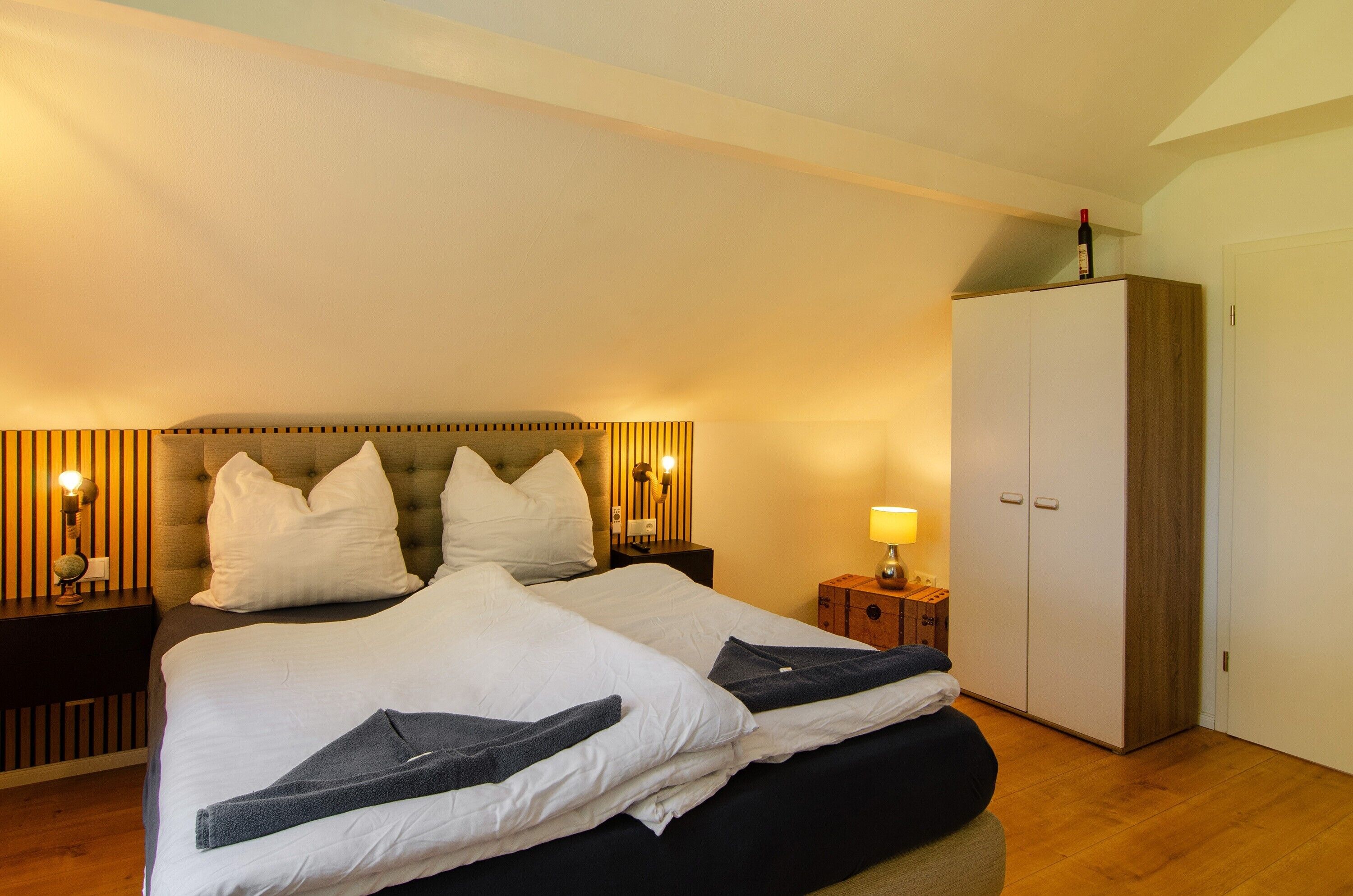 A hotel room with a large bed, two bedside tables with lamps, a wardrobe, and a wooden floor.