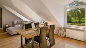 Ein modernes Schlafzimmer im Loft-Stil mit schrägem Dach, einer Essecke mit Holztisch und -stühlen sowie einem großen Fenster mit Blick auf Grünflächen.