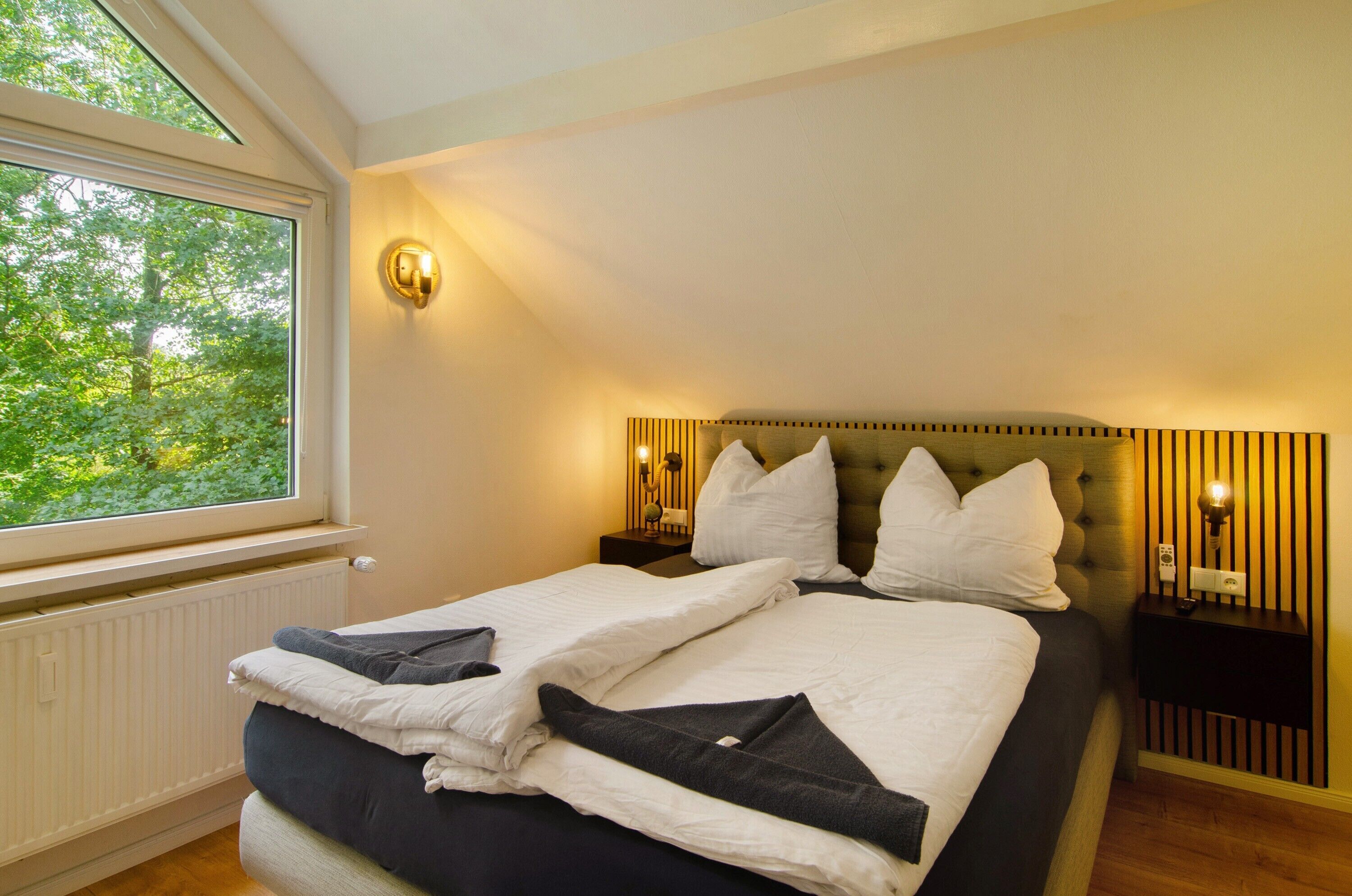 Ein Schlafzimmer mit einem großen Fenster, einem Bett mit weißer Bettwäsche, einem Kopfteil mit vertikalen Lamellen und zwei Nachttischlampen.