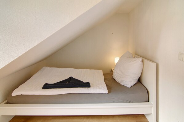 Una cama individual con un colchón blanco y una manta gris, una almohada blanca y una prenda oscura doblada sobre la cama.