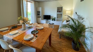 A dining area with a wooden table set for four, a sofa, a TV, and a bookshelf.