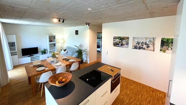 A modern kitchen with a central island, dining table, and chairs. There is a television, bookshelf, and artwork on the walls.