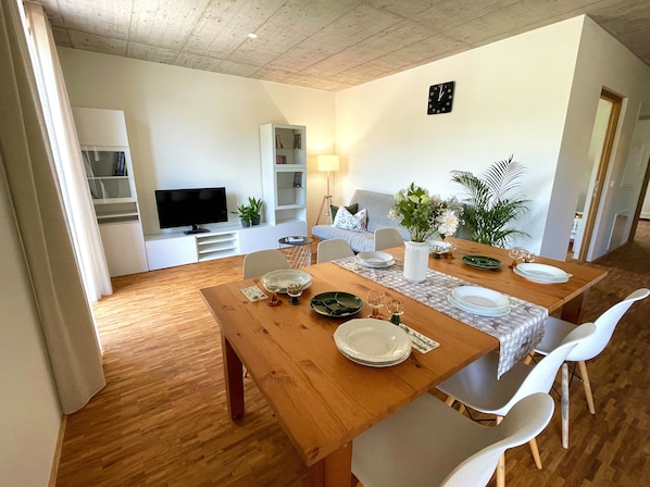 A dining area with a wooden table set for four, surrounded by white chairs. A television is mounted on a white cabinet, and there is a bookshelf with books and decorative items.