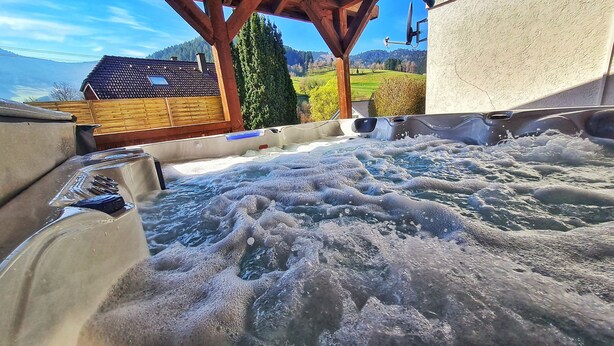 Een hottubgedeelte met een houten terras en uitzicht op een huis en bergen.