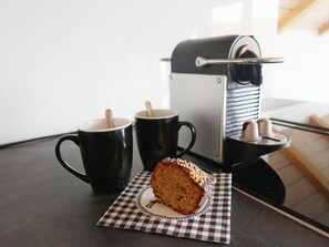 Une part de gâteau sur une serviette à carreaux, deux tasses noires et une machine à café sur une table.
